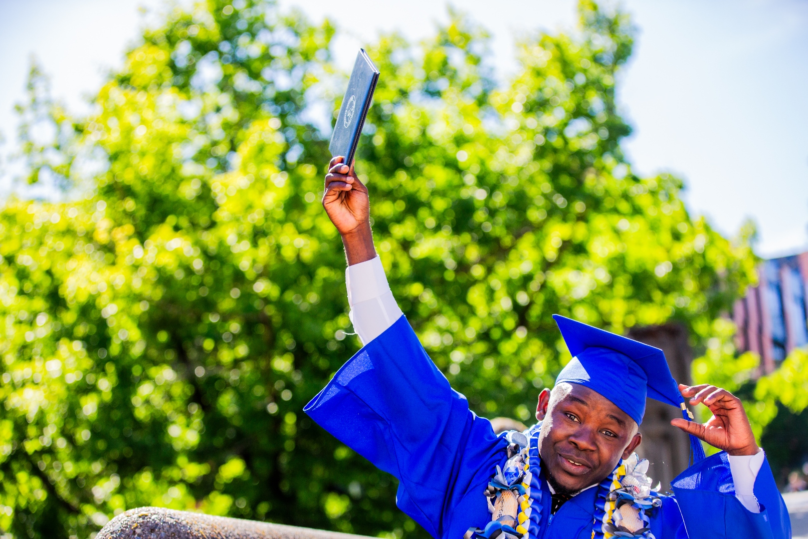 A Seattle Colleges graduate raises his diploma toward the camera under a canopy of summer green.